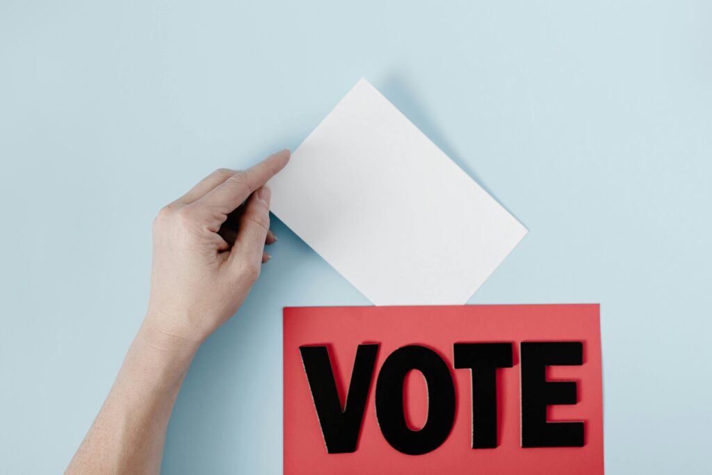pexels-photo-8850718-8850718 A hand places a ballot into a red box with the word VOTE against a blue background.